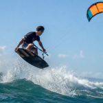 Atleta brasileiro praticando kitesurf em uma onda com vento forte e a pipa no céu