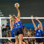 Jogadoras de vôlei feminino disputam uma partida em quadra indoor, com uma atleta saltando para atacar a bola.