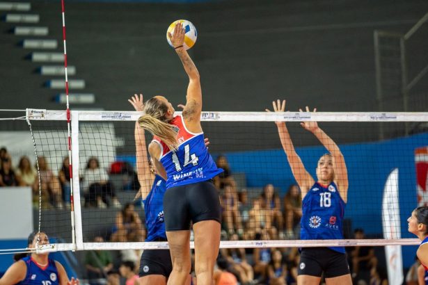 Jogadoras de vôlei feminino disputam uma partida em quadra indoor, com uma atleta saltando para atacar a bola.