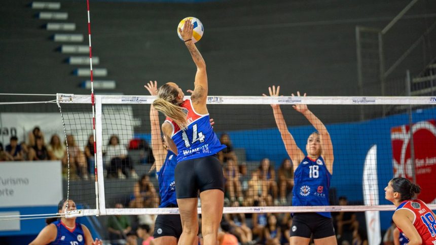 Jogadoras de vôlei feminino disputam uma partida em quadra indoor, com uma atleta saltando para atacar a bola.