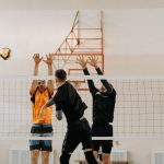 Jogadores disputam uma bola junto à rede durante uma partida masculina de vôlei em quadra coberta.