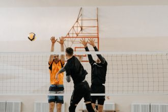 Jogadores disputam uma bola junto à rede durante uma partida masculina de vôlei em quadra coberta.
