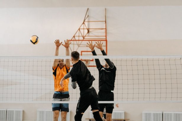 Jogadores disputam uma bola junto à rede durante uma partida masculina de vôlei em quadra coberta.