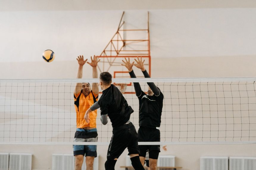 Jogadores disputam uma bola junto à rede durante uma partida masculina de vôlei em quadra coberta.