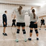 Atletas adultas treinam vôlei em uma quadra coberta, reunidas ao redor da rede.