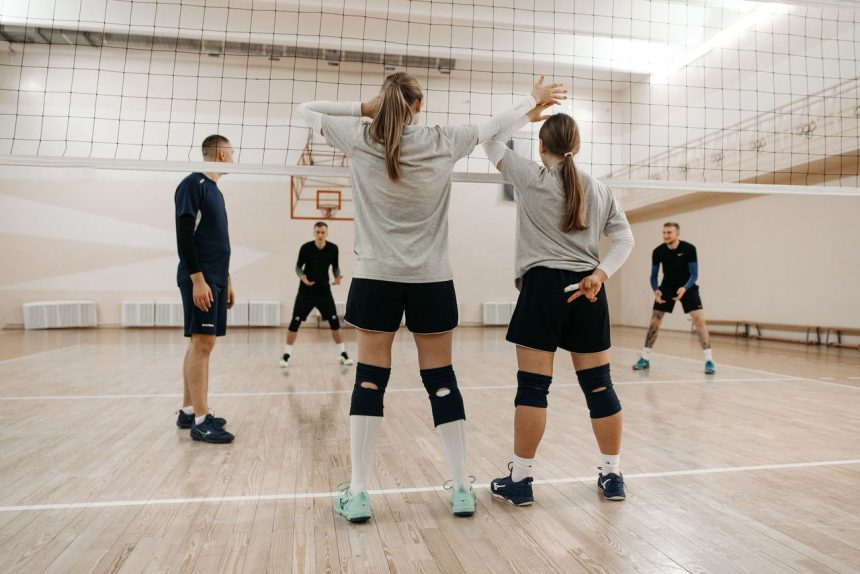 Atletas adultas treinam vôlei em uma quadra coberta, reunidas ao redor da rede.