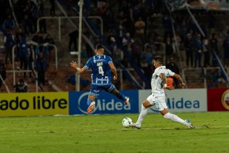 Dois jogadores disputam a bola durante uma partida de futebol noturna, diante de torcedores.