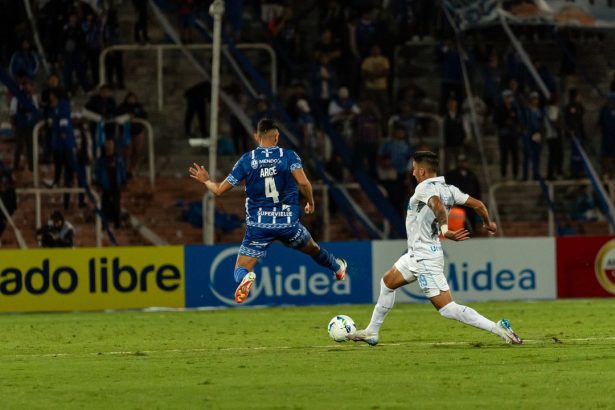 Dois jogadores disputam a bola durante uma partida de futebol noturna, diante de torcedores.