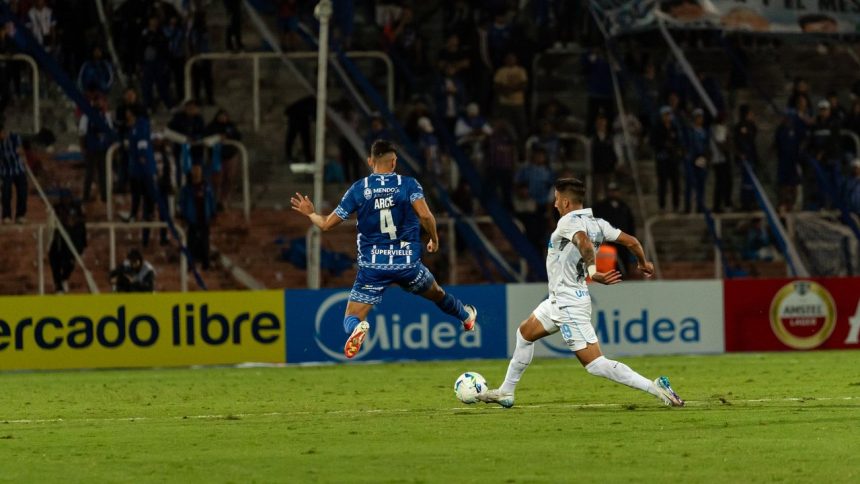 Dois jogadores disputam a bola durante uma partida de futebol noturna, diante de torcedores.