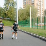 Duas jogadoras de futebol caminham próximas ao campo em um dia ensolarado.