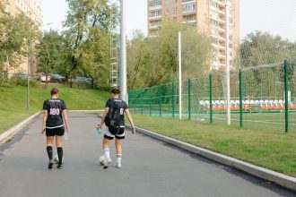 Duas jogadoras de futebol caminham próximas ao campo em um dia ensolarado.